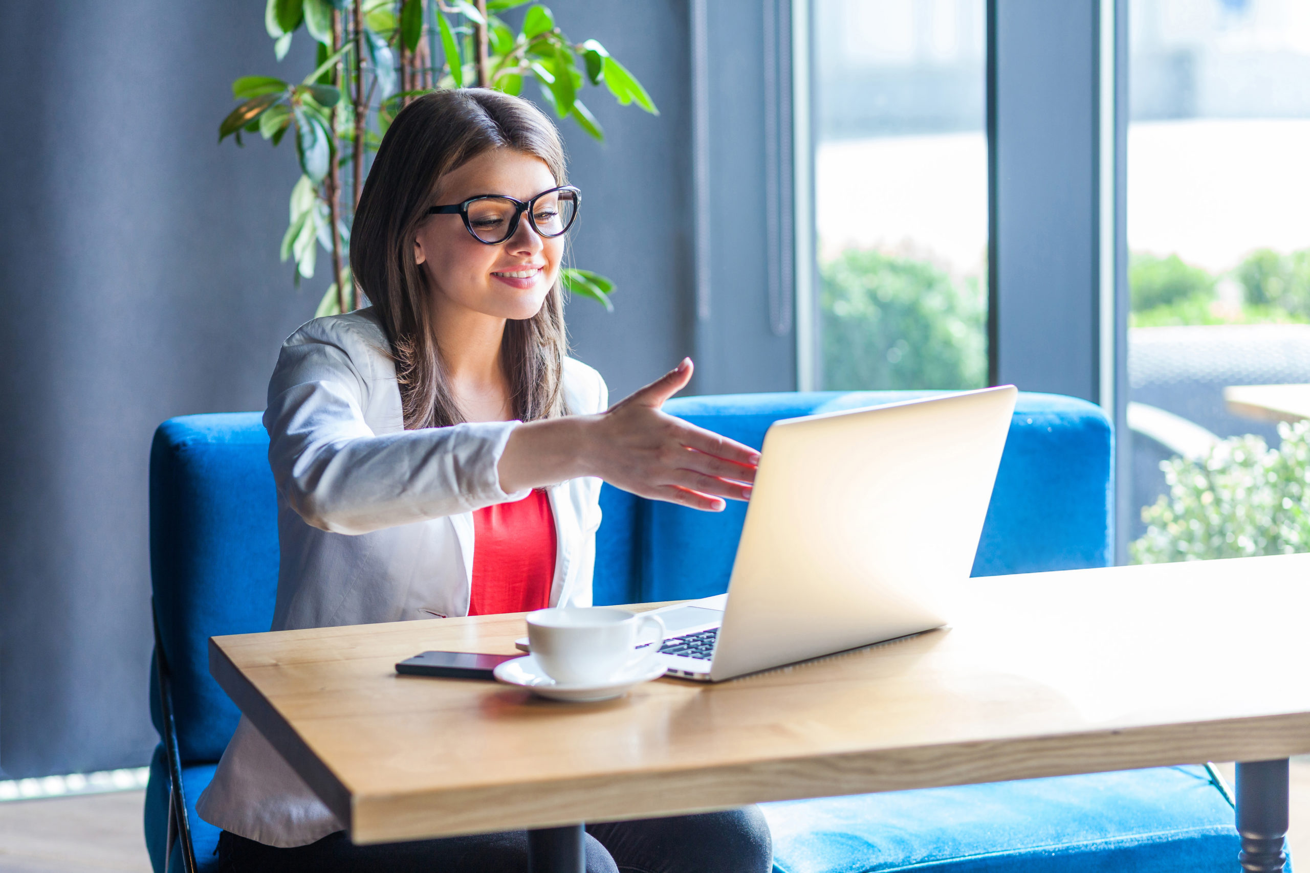 Portrait of happy beautiful stylish young woman in glasses sitting, looking at her laptop screen on video call and giving hand to handshake, toothy smile. indoor studio shot, cafe, office background. Using The Client Portal
